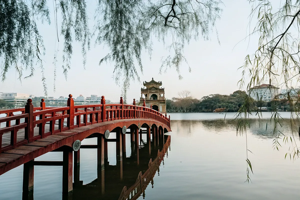 A red wooden bridge over a calm lake in Hanoi, framed by willow branches
